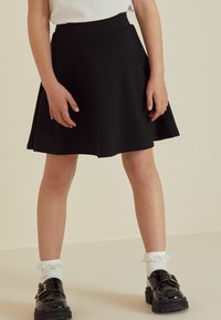 Schwarzer, ausgestellter Rock mit hoher Taille, aus einem weichen Stoff. Kombiniert mit schwarzen Lackschuhen und weißen Rüschen-Socken.