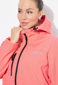 Koralfarvet softshell-jakke med lynlås foran, høj hals med hætte og sorte lynlåse. "HYRAR" logo i lyseblå på brystet.