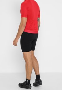 Man wearing a red short-sleeve cycling jersey, black padded shorts, black socks, and black cycling shoes, standing against a white background.