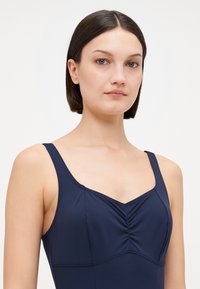 Maillot de bain bleu marine avec un décolleté en V et des détails froncés au niveau de la poitrine. Fabriqué dans un tissu lisse et extensible. Équipé de fines bretelles.