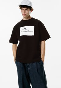 Sort t-shirt med kortærmer og en hvid grafik af en flue på fronten. Bæres med løstsiddende blå denimbukser.