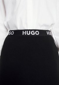 Détail de la taille d'une jupe noire avec une ceinture élastique de marque portant l'inscription "HUGO", portée avec une chemise blanche rentrée.