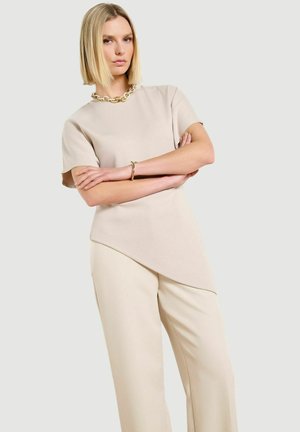 Donna con capelli biondi lisci con le braccia incrociate, indossa un top beige asimmetrico a maniche corte, pantaloni a gamba larga coordinati, collana a catena dorata e bracciale.