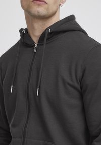 Zwarte rits-hoodie gemaakt van zacht katoen. Voorzien van een trekkoord met metalen punten en een voorzak. Eenvoudig, getailleerd ontwerp met een capuchon.