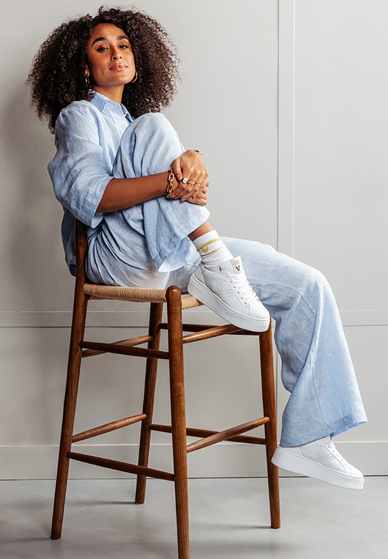 Weiße Sneakers mit strukturierten Sohlen, ein hellblaues Leinen-Outfit und ein Holzstuhl, der eine entspannte Sitzpose vor einem neutralen Hintergrund zeigt.