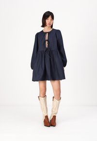 Vestido de denim azul-marinho com decote amarrado, mangas bufantes e cintura franzida. Combinado com botas de cowboy castanhas e creme. Textura suave.