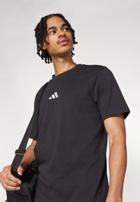Zwarte katoenen T-shirt met korte mouwen, voorzien van een klein wit Adidas-logo op de borst. Het model houdt een zwarte tas met een strap vast.