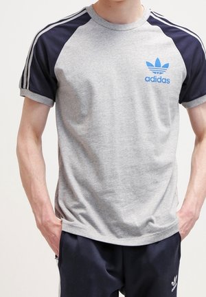 Man draagt een grijs Adidas T-shirt met marineblauwe mouwen en bijpassende marineblauwe Adidas broek, staat met handen in de zakken.