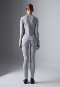 Persona in piedi di spalle, che indossa un aderente top grigio chiaro a maniche lunghe e leggings, con calzini neri, su uno sfondo grigio semplice.