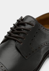 Svart skinnsko med brune lisser, perforert design på oversiden og en jevn tekstur. Sålen har en subtil kurve ved kanten.