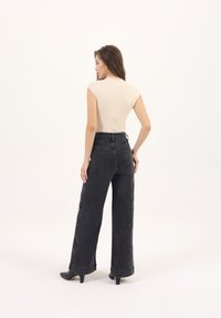 Top sin mangas de canalé beige combinado con vaqueros negros de pierna ancha y cintura alta. Los vaqueros tienen bolsillos traseros y un dobladillo enrollado; la modelo lleva unos tacones negros.
