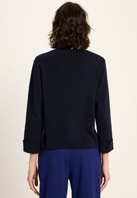 Frau mit schulterlangem, lockigem Haar, trägt ein schwarzes, langärmliges Oberteil und blaue Hosen, steht mit dem Rücken zu einem hellen Hintergrund.