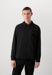 Black zip-up shirt with a textured fabric, pointed collar, long sleeves, and a chest pocket detail. Worn over dark pants.