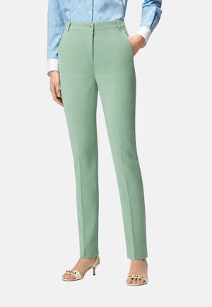 Donna che indossa pantaloni verde menta slim fit, camicia a righe blu e sandali bianchi con tacco e borchie dorate, in piedi con una mano in tasca.