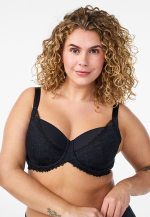 Ženska s kodrastimi lasmi, ki nosi črni čipkasti nedrček z žico, se smeji, na podlahti ima vidno tetovažo, na enostavni beli podlagi.