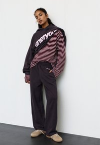 Hanorac închis la culoare cu logo roz, purtat peste un tricou cu mâneci lungi în dungi bordo și albe, asortat cu pantaloni negri largi și pantofi bej.