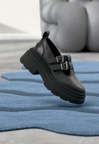 Schwarze Lederschuhe mit klobigen Plateausohlen und zwei Metall-Schnallenakzenten; verfügt über eine abgerundete Spitze und eine glatte Textur.