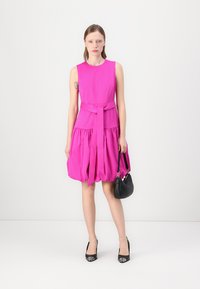 Fuchsiové šaty bez rukávů s páskem v oblasti pasu a rozšířenou, nařasenou sukni. V kombinaci s malou černou kabelkou a černými lodičkami.