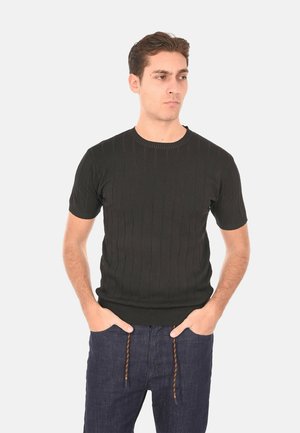 Uomo che indossa una maglietta nera a maniche corte a costine e pantaloni blu scuro con coulisse marroni, mani in tasca, guarda leggermente verso destra.