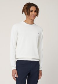 Witte lange mouwen trui met geribbelde boorden en zoom, met een klein grijs logo op de borst. Gedragen over donkere marineblauwe broek.