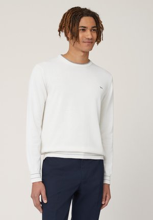 Witte lange mouwen trui met geribbelde boorden en zoom, met een klein grijs logo op de borst. Gedragen over donkere marineblauwe broek.