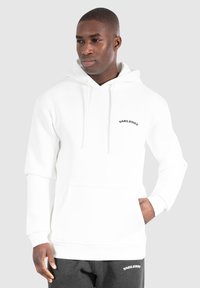 Haut à capuche blanc avec cordon de serrage, poche avant et logo texte noir "SMILODON" sur la poitrine. Porté avec un pantalon de survêtement gris.