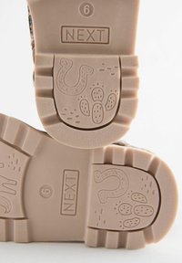 Semelles de chaussures en caoutchouc beige clair avec une texture antidérapante. Présente un logo "NEXT" et des motifs ludiques de traces de pattes sur le profil. Taille 6 indiquée.