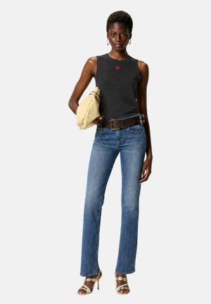 Donna che indossa un top nero senza maniche con un cuore rosso, jeans blu, sandali beige con tacco alto, cintura marrone, tiene in mano una borsa beige.