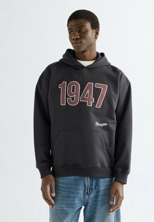 Jonge man die een zwarte hoodie draagt met grote cijfers "1947" en tekst "Wrangler", gecombineerd met een blauwe spijkerbroek, staand tegen een witte achtergrond.