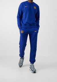 Sudadera y pantalones de chándal de algodón azul con detalles amarillos, con logo de Nike y escudo de Barcelona. Acabado texturizado y ajuste relajado.