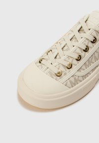 Beige sneaker met een textuurstof bovenkant, voorzien van een subtiel logo patroon, gouden oogjes en een witte rubbere zool.
