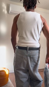 Bărbat cu dreadlocks, îmbrăcat într-un maiou alb și pantaloni gri, stă în interior ținând un obiect înfășurat în plastic lângă un aparat de aer condiționat de pe perete.