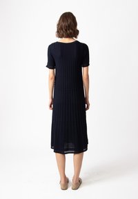 Haljina do koljena, tamnoplave boje, s kratkim rukavima, rebrastom teksturom, okruglim izrezom i bočnim prorezima; završena s suptilnim otvorenim šavnim detaljima.