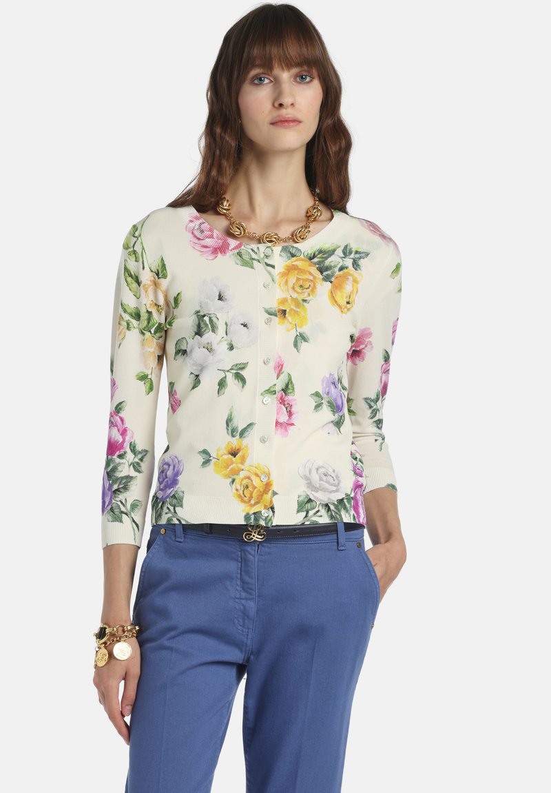 Virágos krémszínű kardigán színes rózsákkal; háromnegyedes ujjú; gombos záródású; kék nadrággal és arany nyaklánccal viselve.
