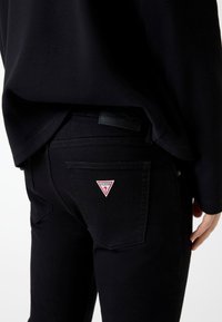 Czarne dżinsy denim o wąskim kroju. Posiadają tylnią kieszeń z czerwonym, trójkątnym naszywką z logo "GUESS" oraz teksturowany czarny pas na talię.