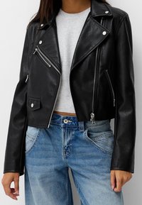 Czarna skórzana kurtka motocyklowa o kroju cropped, wykonana z ekologicznej skóry, z metalowymi suwakami i zatrzaskami, zestawiona z szarym swetrem o prążkowanej strukturze i niebieskimi dżinsami.