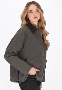 Junge Frau mit langen, welligen Haaren, die eine dunkelgrüne, geknöpfte Jacke mit Cordkragen, die Hand in der Tasche, und schwarze Jeans trägt.