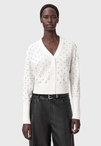 Eine Frau trägt einen weißen, knöpfbaren Cardigan mit silbernen Sternmustern und schwarze Lederhosen mit einem schwarzen Gürtel, während sie vor einem schlichten Hintergrund steht.