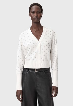 Kvinde iført en hvid knaplukket cardigan med sølvstjernesymbole og sorte læderbukser med et sort bælte, stående mod en ensfarvet baggrund.