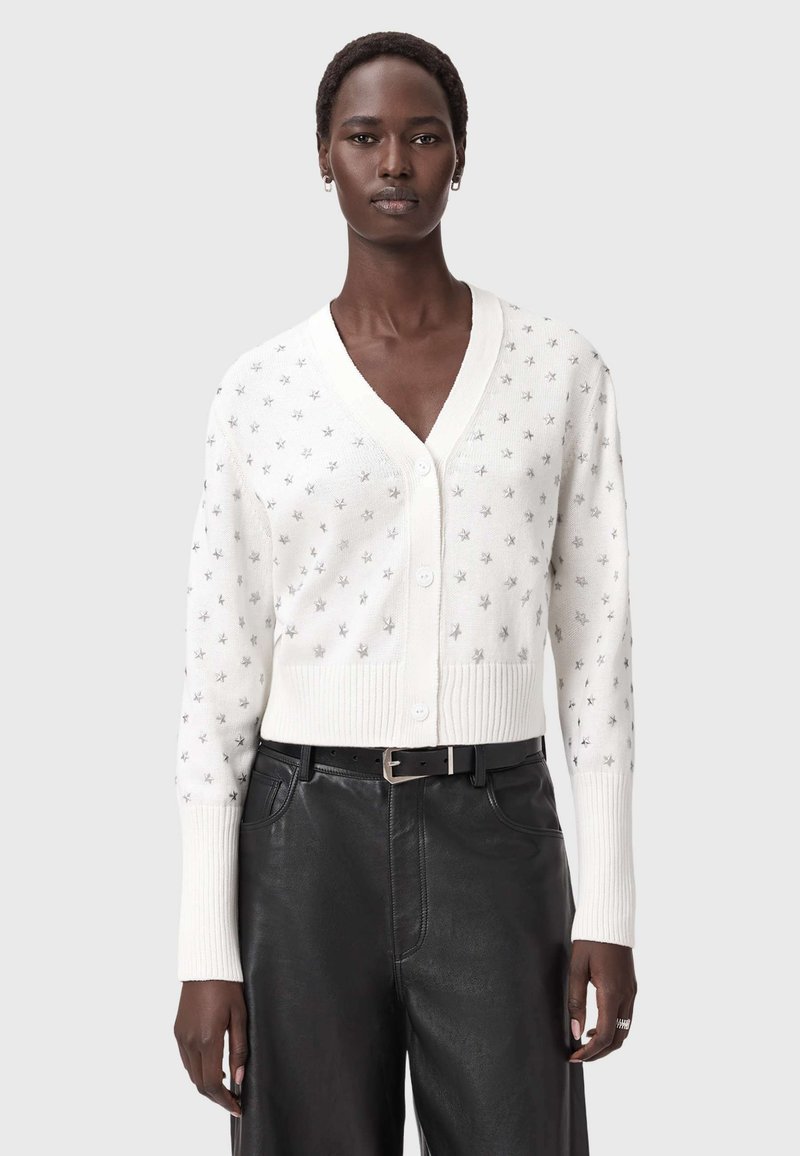 Eine Frau trägt einen weißen, knöpfbaren Cardigan mit silbernen Sternmustern und schwarze Lederhosen mit einem schwarzen Gürtel, während sie vor einem schlichten Hintergrund steht.