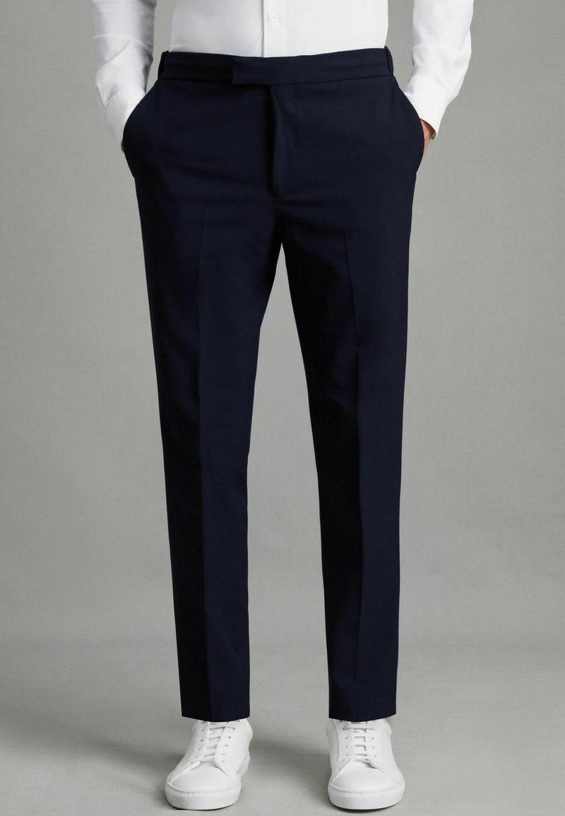 I pantaloni tailored blu navy presentano una linea frontale piatta, due tasche laterali e una vestibilità affusolata. Abbinati a sneakers bianche, creano un look moderno.
