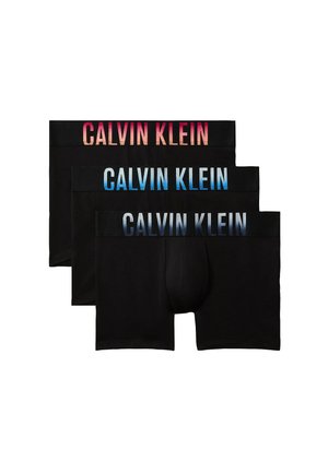Hombre musculoso de pie descalzo sobre fondo gris, vistiendo calzoncillos bóxer negros Calvin Klein con logo azul en la cinturilla.
