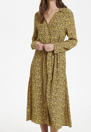 Žena v žlutých midi šatech s černým květinovým potiskem, dlouhými rukávy, límcem a zapínáním na knoflíky vpředu, ruce sepnuté v pase.