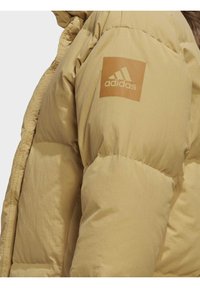 Světle žlutá péřová bunda s hladkým povrchem a elastickými manžetami. Obsahuje vyražené logo Adidas na levém rukávu pro branding.