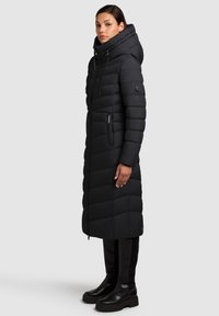 Giacca lunga imbottita nera con cappuccio, caratterizzata da un design trapuntato, tasche laterali con cerniera e silhouette aderente. Texture liscia e materiale caldo.