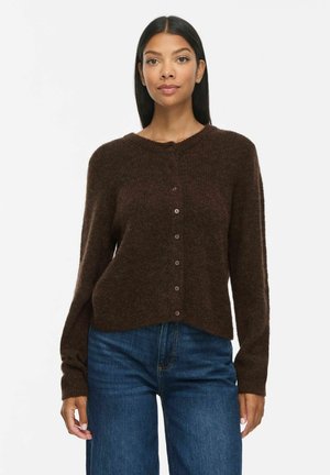 Femme portant un cardigan marron foncé boutonné et un jean bleu, debout devant un fond blanc uni.