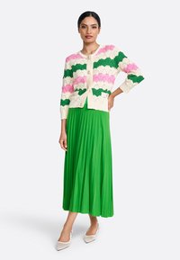 Cardigan en tricot avec un motif festonné rose, vert et crème ; associé à une jupe midi plissée verte et des chaussures à bouts pointus de couleur claire.
