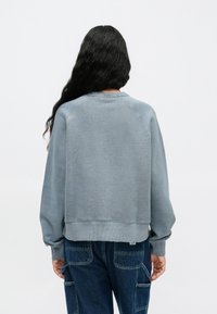 Sudadera azul claro con cuello redondo acanalado y hombros caídos, presenta una textura en la tela y un ajuste relajado, combinada con jeans oscuros.