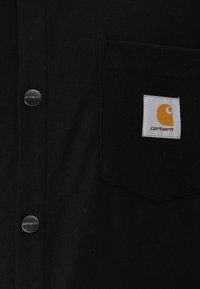 Zwarte flanellen shirt met zilveren drukknoopsluitingen, een borstzak en een oranje en witte Carhartt-logo patch. Zachte textuur, recht gesneden ontwerp.
