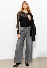 Haut noir en dentelle à manches longues avec un col transparent, associé à un jean gris taille haute à jambe large et des bottines noires à talons.